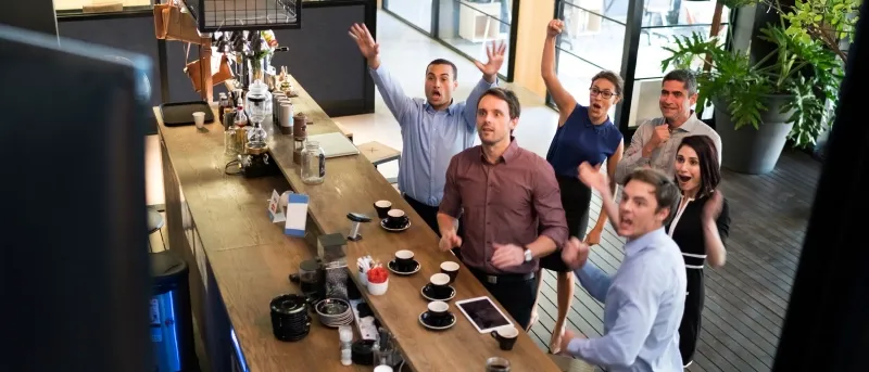 A group of six diverse office workers in a modern break room stand behind a counter, cheering and raising their arms in excitement while watching something off-camera.